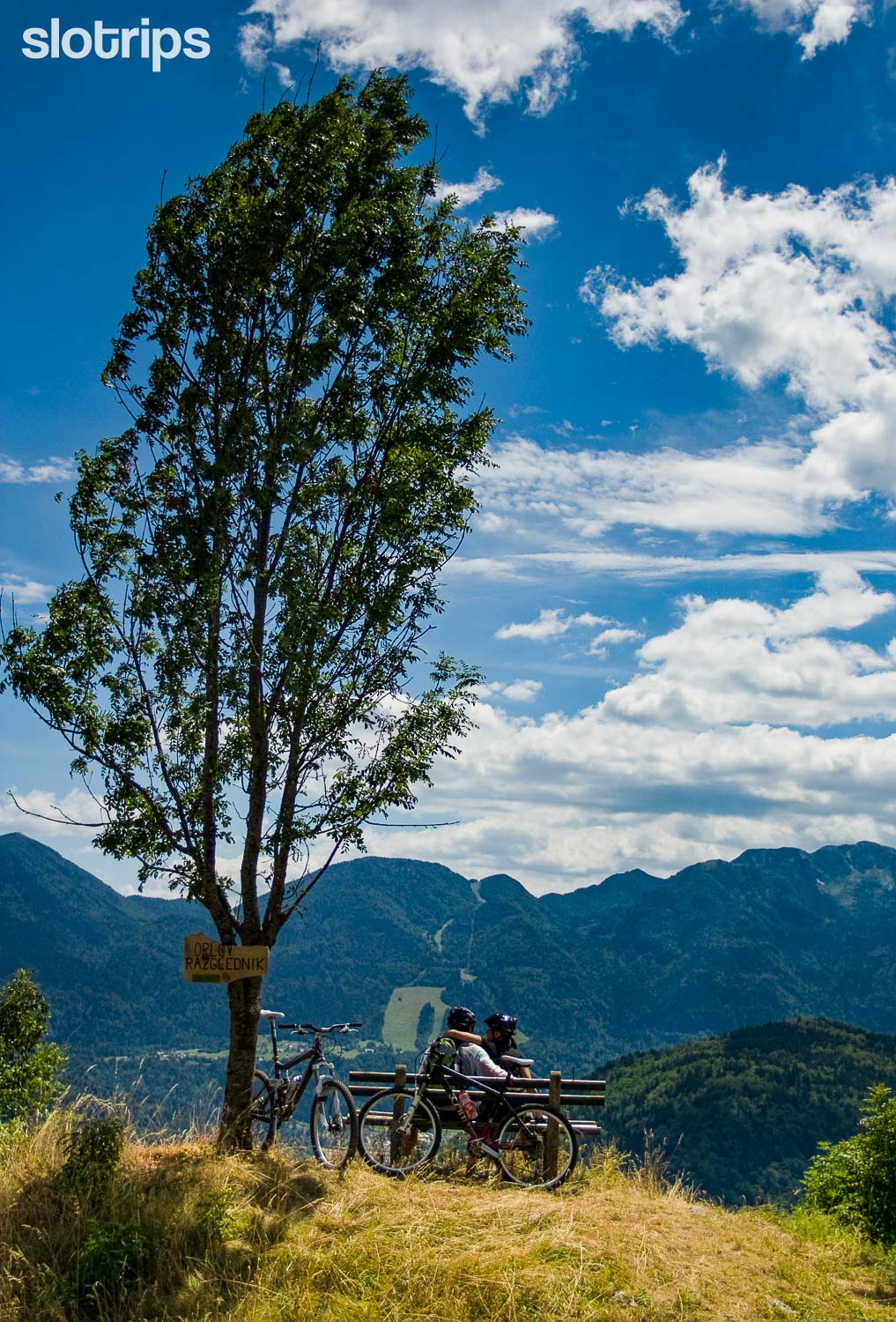 A couple on a mountain biking trip taking a rest in the alpine landscape at Orlov Razglednik in Julian Alps, Slovenia, overlooking Bohinjska Bistrica and former Kobla Ski Resort
