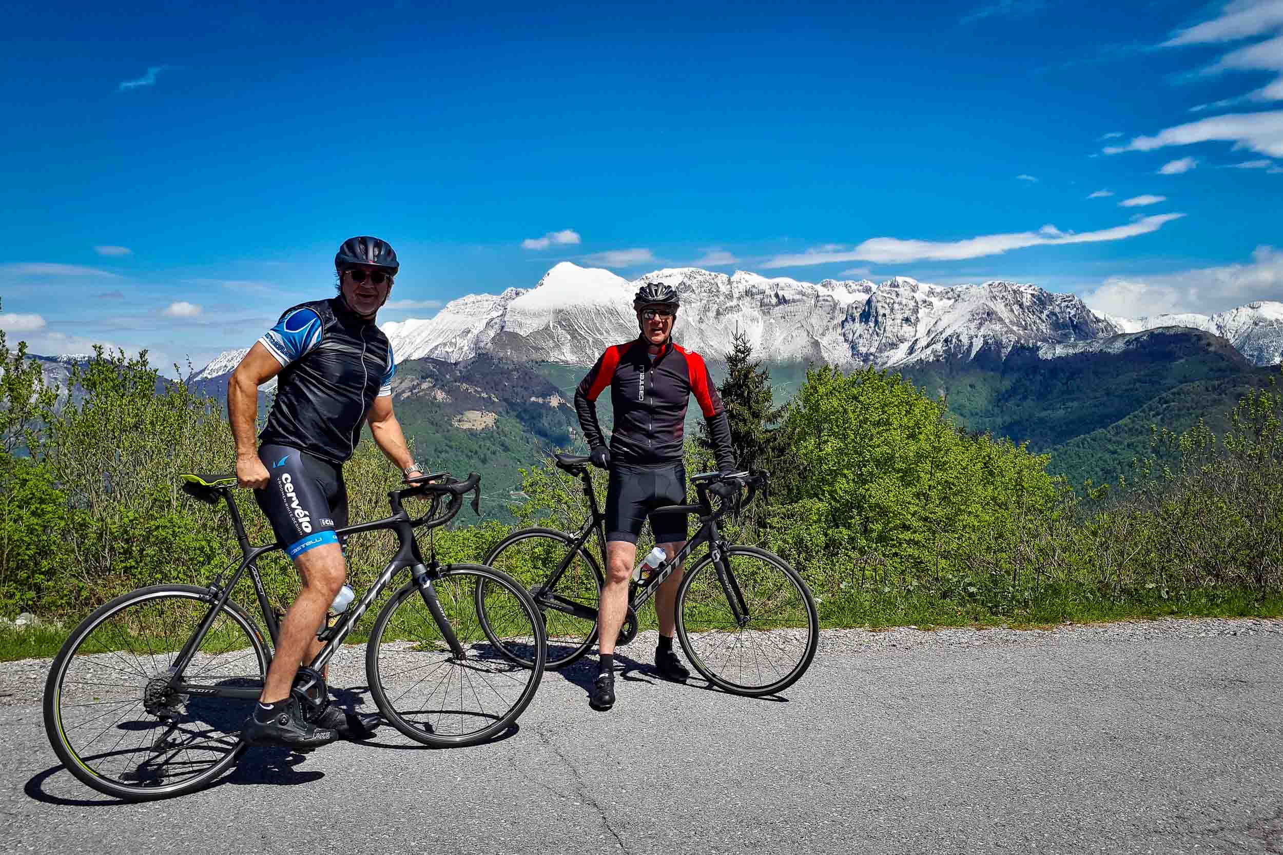 Two road cyclists on a quiet asphalt road biking from the Soca Valley to the Goriska Brda wine region in Slovenia with snowcapped Mt. Krn in the background