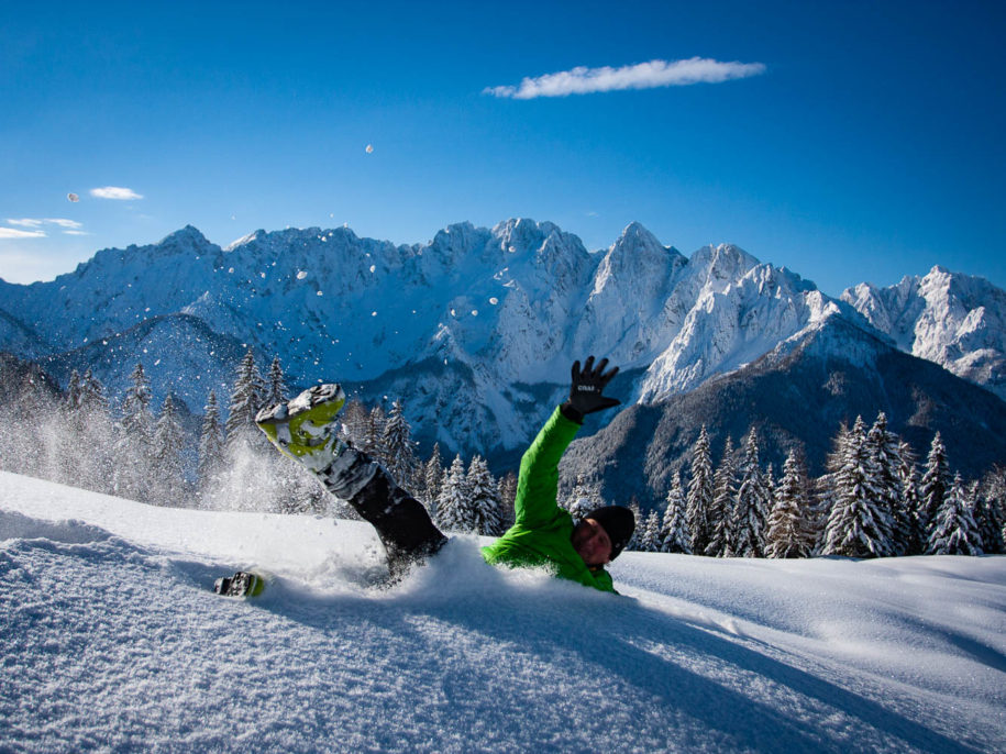 Back country skier joyfully throws himself in deep powder snow backed with a beautiful snow covered mountain panorama of Julian Alps.
