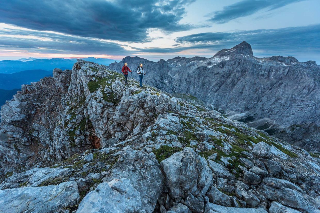 A couple trekking along a stony ridge next to a spectacular limestone massif of the Julian Alps after sunset.
