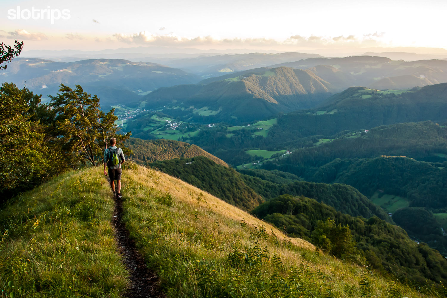 Hiker descending the grassy Blegos mountain ridge in the sunset, surrounded with the wooded Skofja Loka hills.