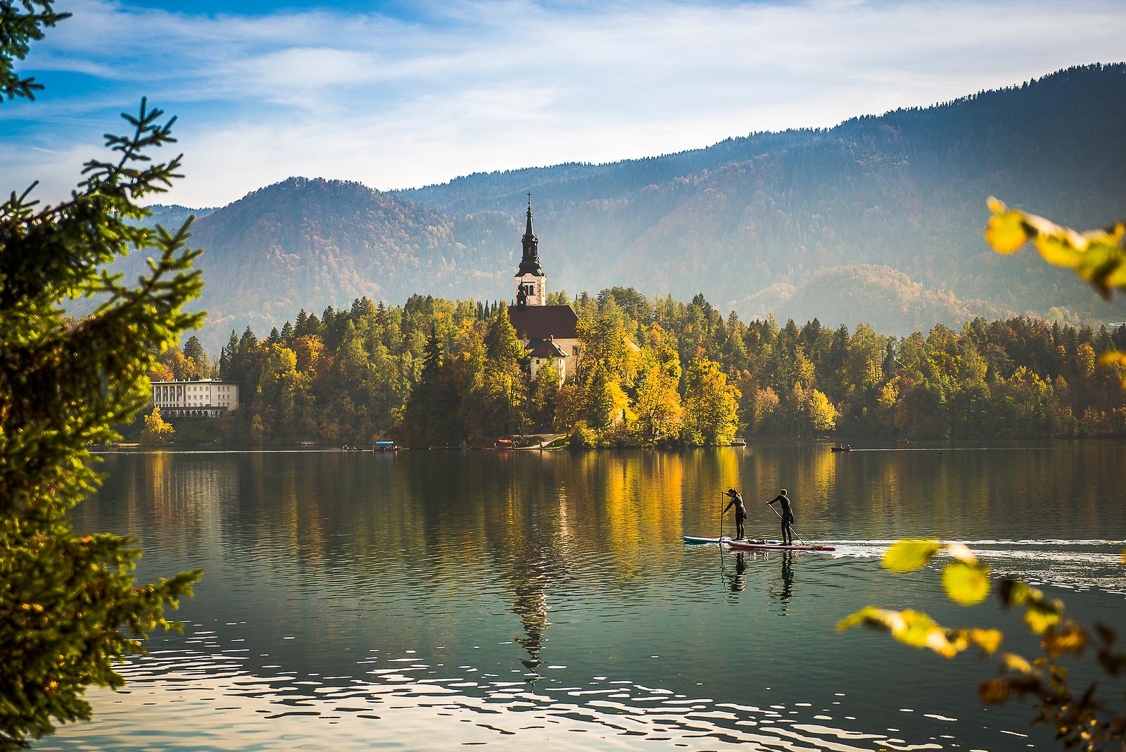 Lake Bled with a couple stand up paddling in the foreground and an island with a Church of the Mother of God on the Lake and former president Tito's villa in the background