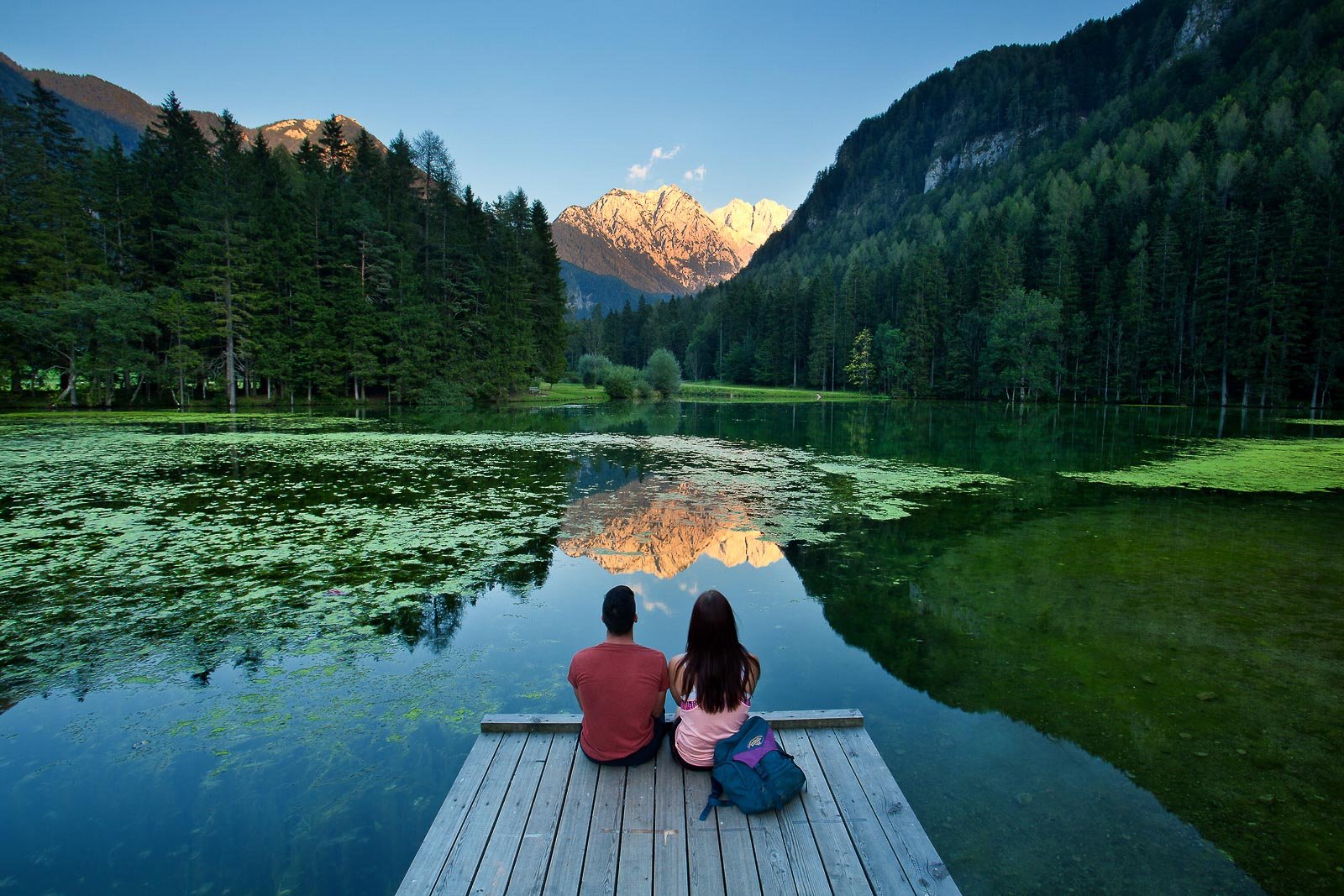 A couple sitting on a pier at Plansarsko jezero lake in Jezersko, looking at the highest peaks of the Kamnik-Savinja Alps during a sunset