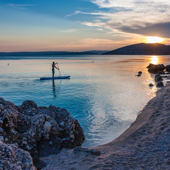 A man on a stand up paddle with the sun setting on the horizon at Krk island in Croatia
